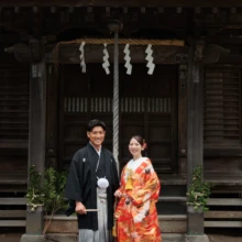 鎌倉 和装 I様・Y様 2026.07.05 鎌倉の古民家に隣接する神社の社殿前で、黒紋付袴の新郎と赤い色打掛の新婦が並ぶ和装前撮り